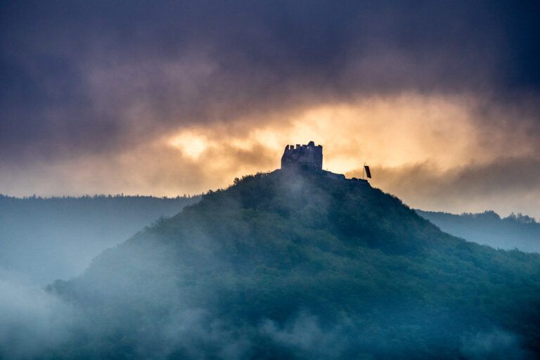 Burgruine auf einem nebelverhangenen H&uuml;gel, dramatische Wolken und Lichtschein am Horizont