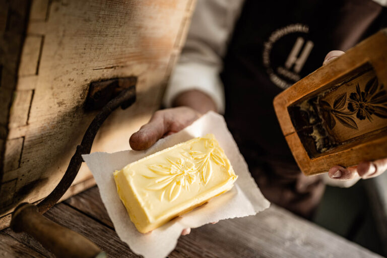 Hand h&auml;lt Butterst&uuml;ck mit Blattpr&auml;gung auf Papier, davor dunkle Sch&uuml;rze mit eingesticktem 'H'-Logo