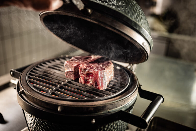 Zwei dicke rohe Steaks auf dem Rost eines ge&ouml;ffneten Keramikgrills, Glut und ge&ouml;ffneter Deckel im Hintergrund