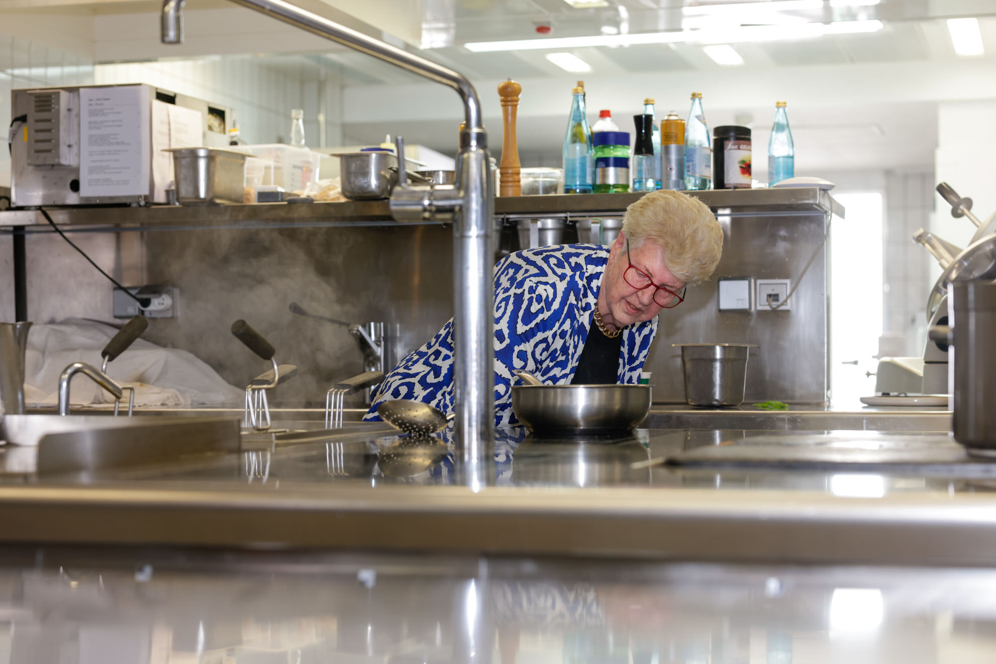 &Auml;ltere Person mit kurzen wei&szlig;en Haaren beugt sich &uuml;ber einen Kochtopf in einer professionellen Edelstahlk&uuml;che mit Gew&uuml;rzen.