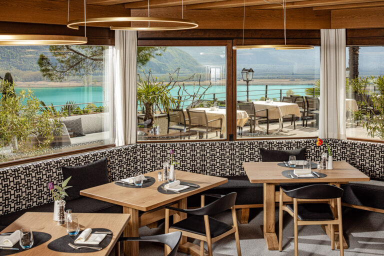 Hotelrestaurant mit Holztischen, gepolsterten Sitzb&auml;nken, gro&szlig;en Panoramafenstern und Terrasse mit Blick auf Kalterer See.