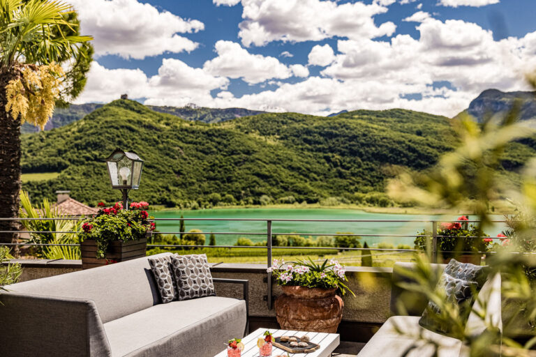 Terrassenbereich mit Sitzbank, Pflanzk&auml;sten und Blick auf den Kalterer See, gr&uuml;ner Hang und wolkiger Himmel.