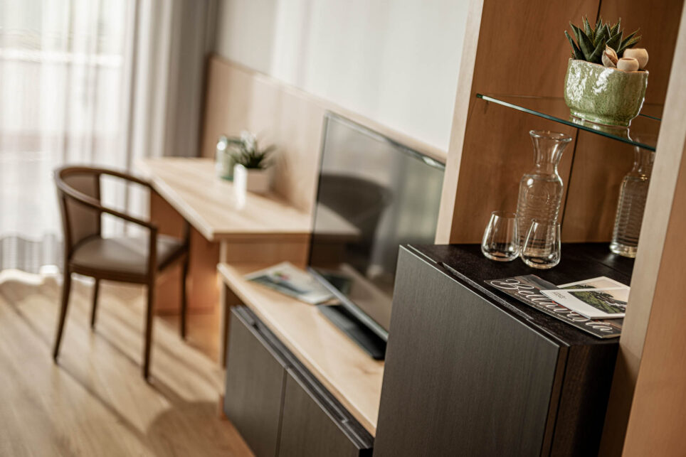 Helles Zimmer mit Schreibtisch und Stuhl, kleine Pflanze, Flachbildschirm auf Kommode, Gläser auf Schrank, Vorhang am Fenster