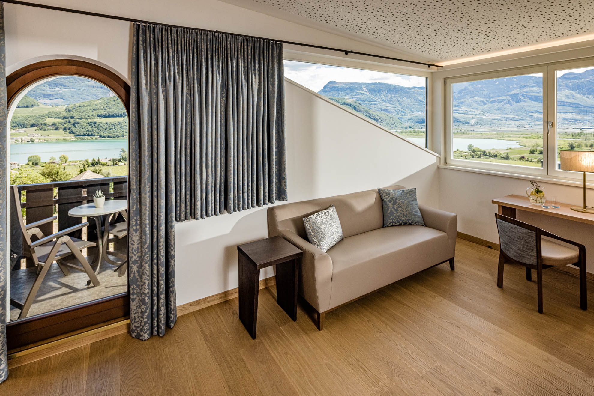 Sofa, Vorhang und Beistelltisch vor gro&szlig;en Fenstern mit Tal- und Bergblick; Holzboden und kleiner Balkon.