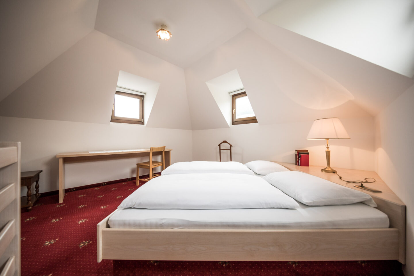 Dachzimmer der Suite mit niedrigem Doppelbett, wei&szlig;er Bettw&auml;sche, zwei Dachfenstern, rotem Teppich und Nachttischlampe.