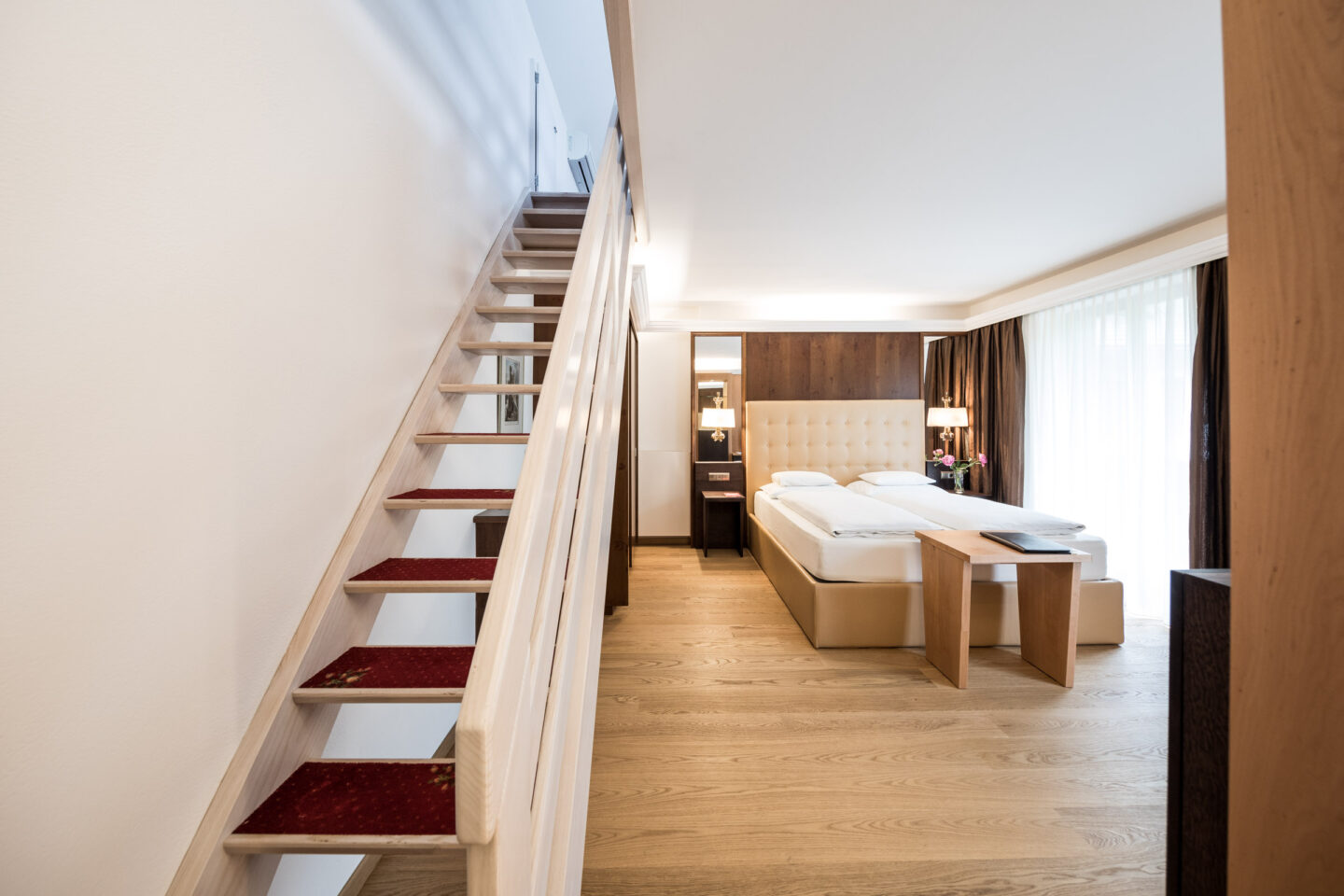 Suitezimmer mit offener Holztreppe links, Doppelbett, Holzfu&szlig;boden, Sitzbank und gro&szlig;en Fenstern mit Vorh&auml;ngen