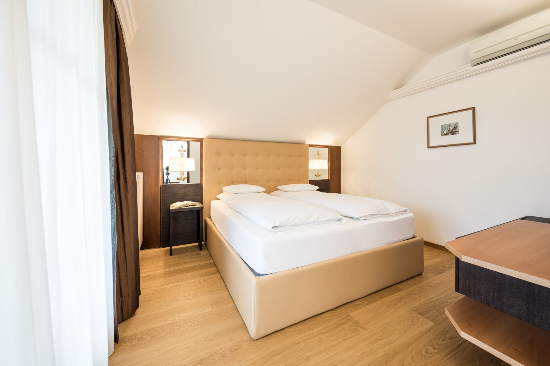 Doppelbett in heller Suite mit schr&auml;ger Decke, zwei Nachttischen mit Lampen, Parkettboden und geschlossenem Vorhang