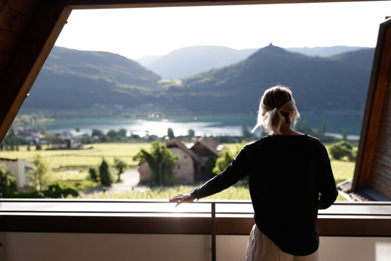 Person lehnt am Balkon und blickt auf den Kalterer See, Uferwiesen und h&uuml;gelige Weinberge im Gegenlicht.