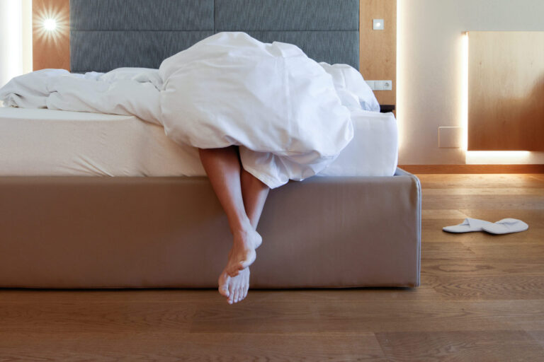 Beine einer Person ragen unter zerw&uuml;hlter wei&szlig;er Bettdecke &uuml;ber die Bettkante in einem Hotelzimmer mit Holzboden.