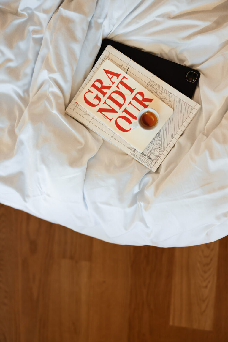 zerknitterte wei&szlig;e Bettdecke auf Holzfu&szlig;boden, Buch 'GRAND CRU' und ein schwarzes Ger&auml;t liegen darauf