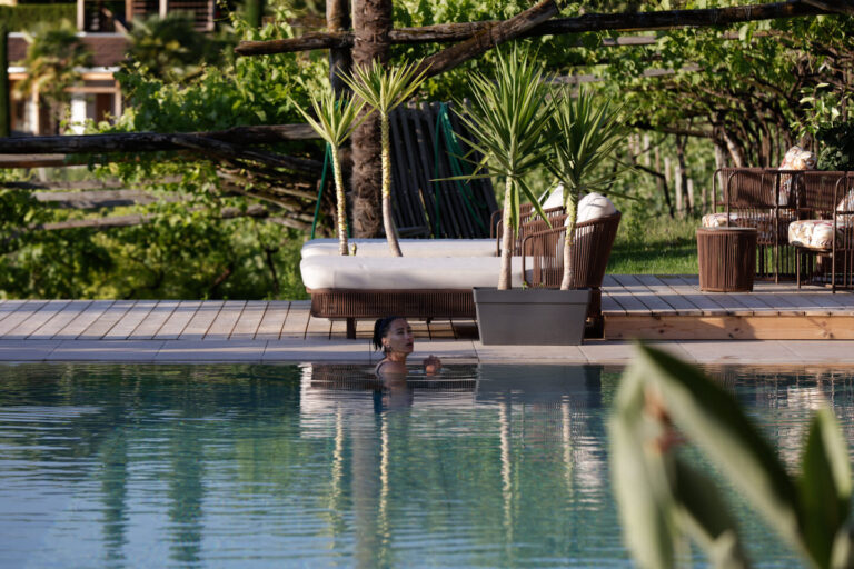Tagesbett mit Kissen und Decke auf Holzdeck am ruhigen Wasser, umgeben von Palmen und Uferbepflanzung