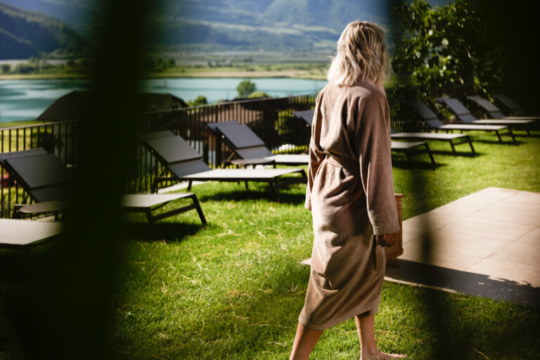 R&uuml;ckenansicht einer Person im Bademantel auf Rasen, geht zu Liegest&uuml;hlen am Ufer; davor dunkle Pflanzen als Rahmen.