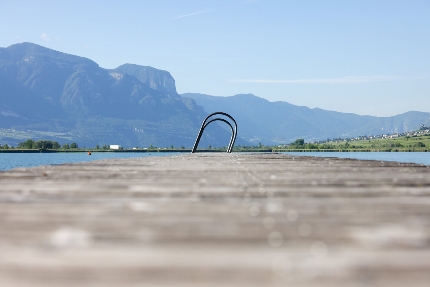 Holzsteg mit gebogener Metallleiter ins Wasser, Schilf am Ufer und Bergkette am Horizont