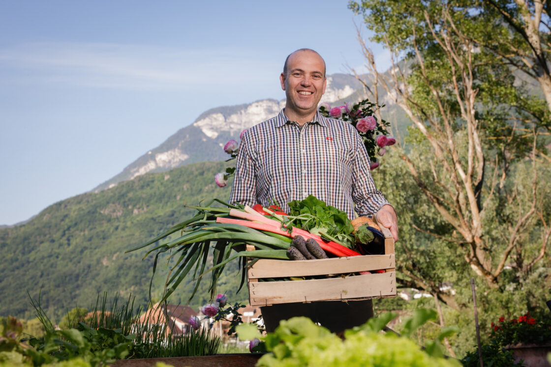 Mann hält Kiste mit Lauch, Karotten, Radieschen und Kräutern, Ernte aus Eigenanbau vor Bergkulisse.