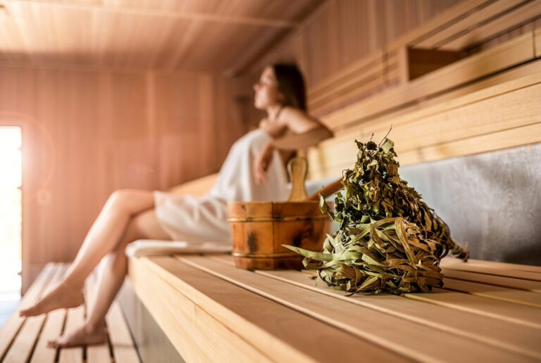 Person mit Handtuch sitzt auf Holzbank in Hotelsauna; Saunak&uuml;bel und Aufgussbesen aus Birkenzweigen im Vordergrund.