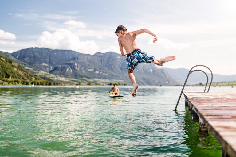 Mann springt von Holzsteg in gr&uuml;nes Wasser des Kalterer Sees; Berge am Horizont, weitere Person auf Steg und Leiter am Ufer.